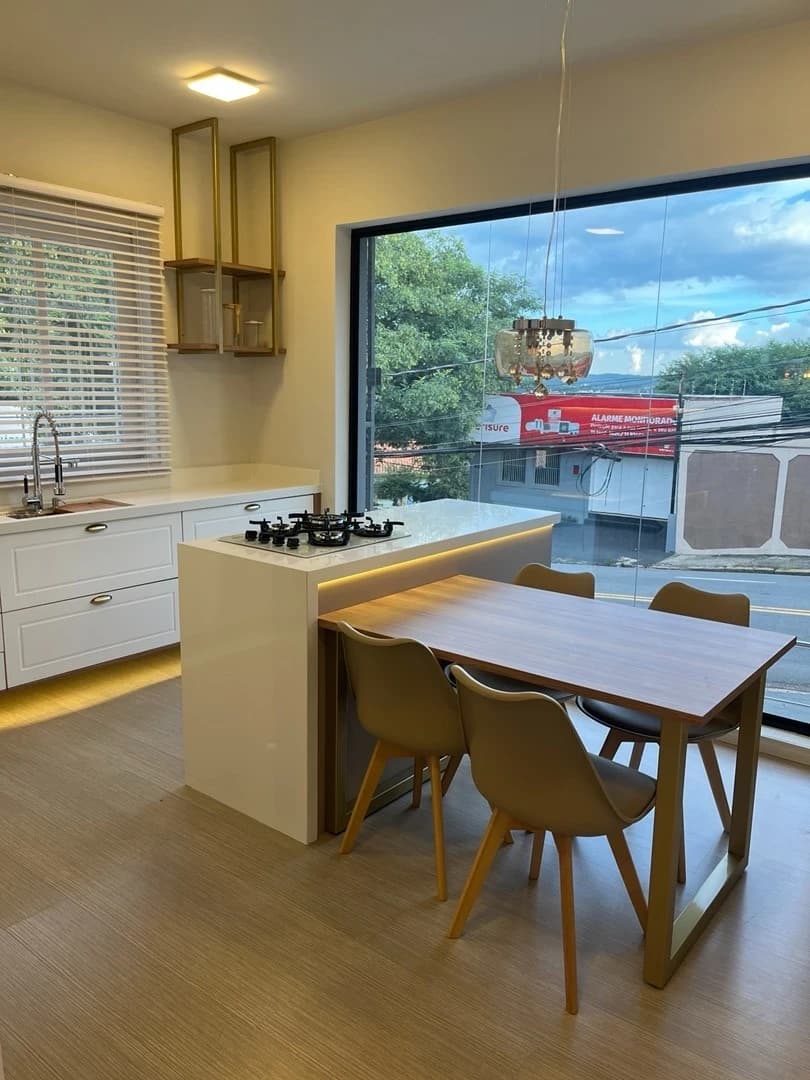 Cozinha Moderna com Vista Ampla: Ambiente moderno e funcional, com linhas clean, armários brancos, bancada em marfim e mesa de madeira. Destaque para a iluminação e a janela que proporciona vista para o exterior.
